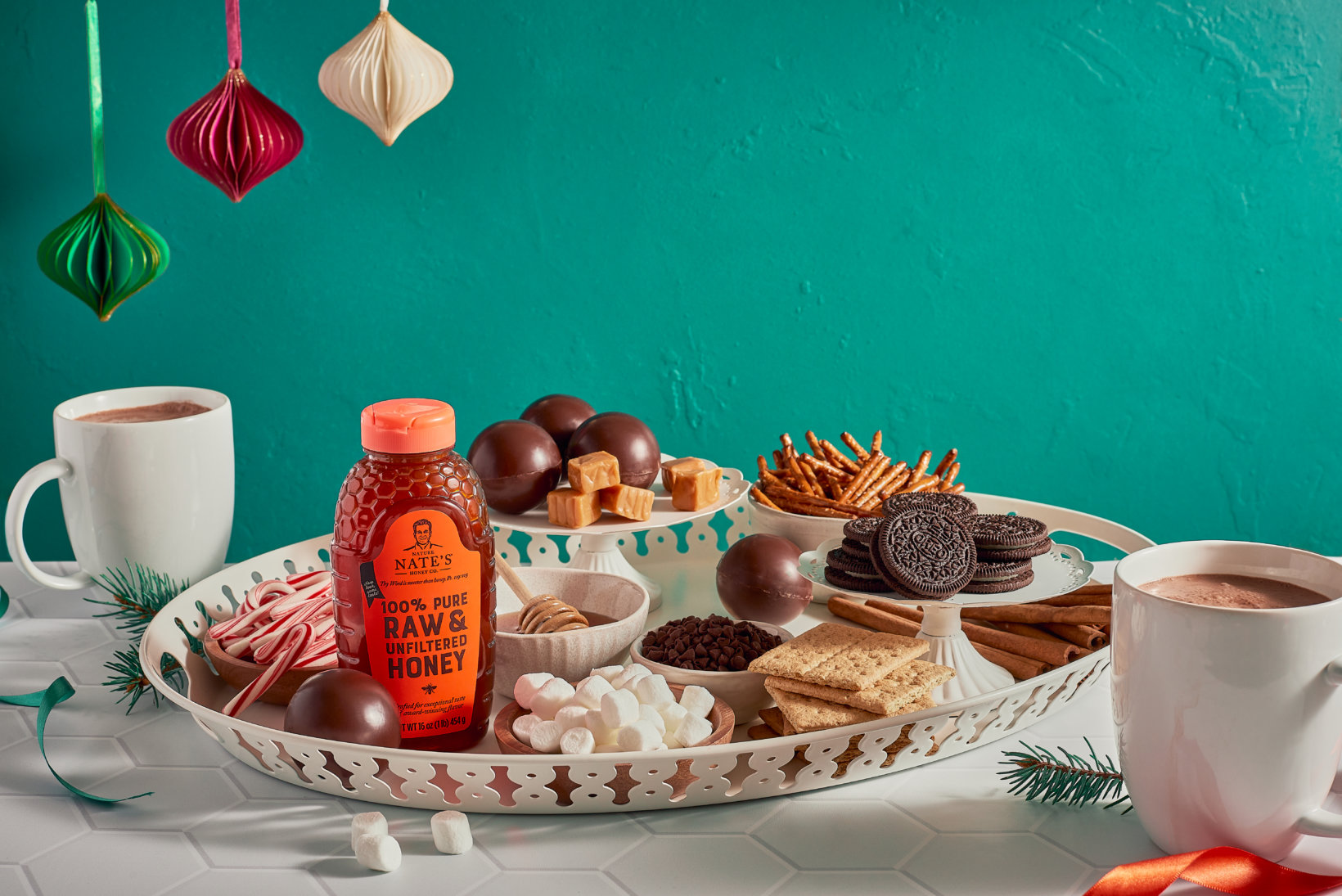 A holiday snack tray with a Nate’s honey bottle, treats, hot chocolate, on a green background with ornaments.