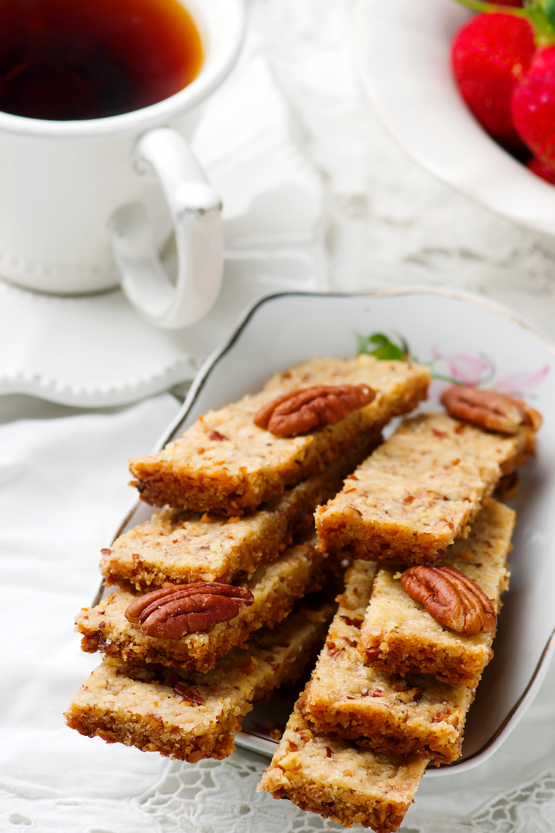 A plate with pecan bars accompanied by a cup of tea and strawberries is placed on a lace doily with a white napkin.