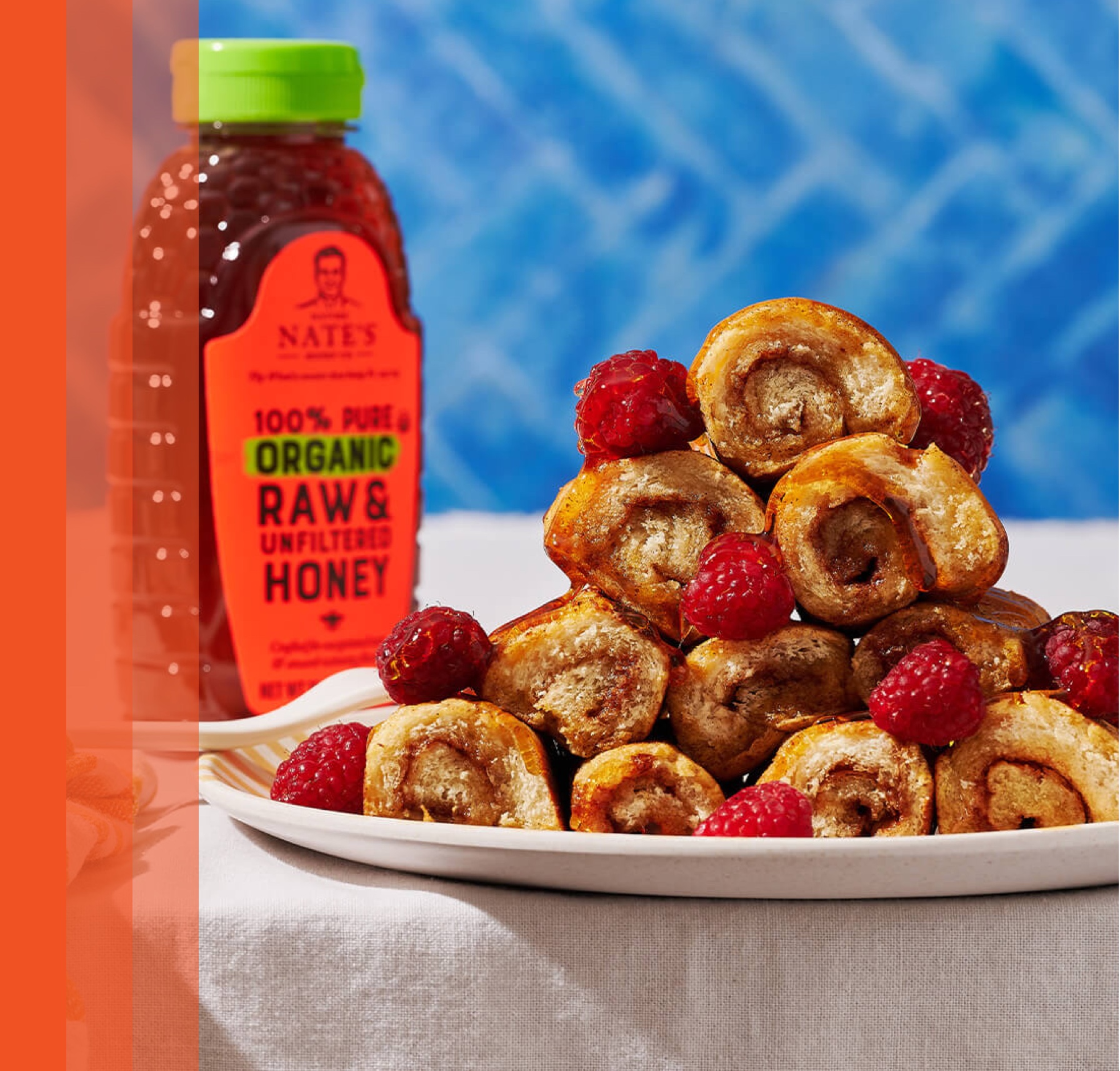 Bottle of Nate's Organic honey next to platter of French toast rollups with raspberries on cloth table and blue background