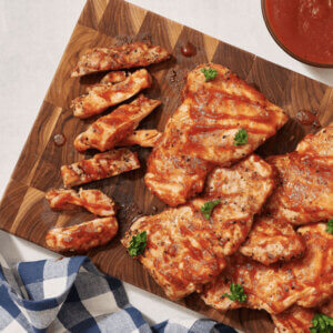 A row of assorted homemade spreads in jars with a bottle of raw honey, against a white brick wall. Grilled chicken with hot honey bbq sauce on wooden platter
