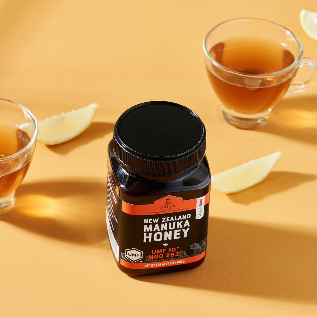A jar of Nate’s New Zealand manuka honey UMF 10+ with cups of tea and lemon slices on a yellow background.