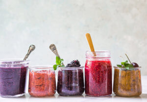 Row of colorful berry jams in glass jars, each with a spoon, against a soft backdrop.