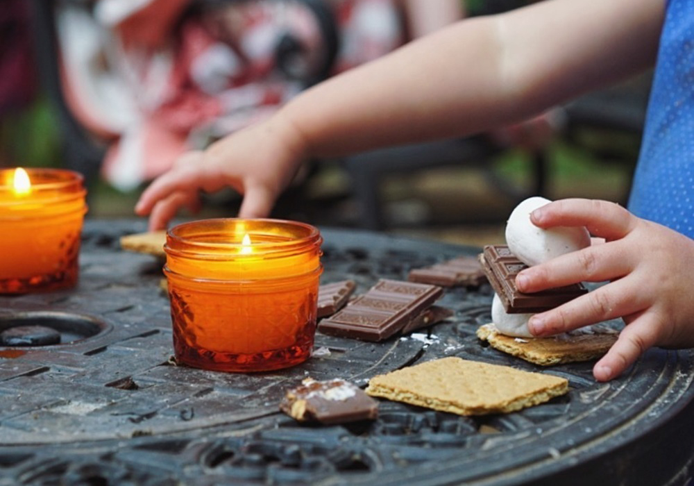 Child's hands preparing s'mores with ingredients and lit candles on an iron table.