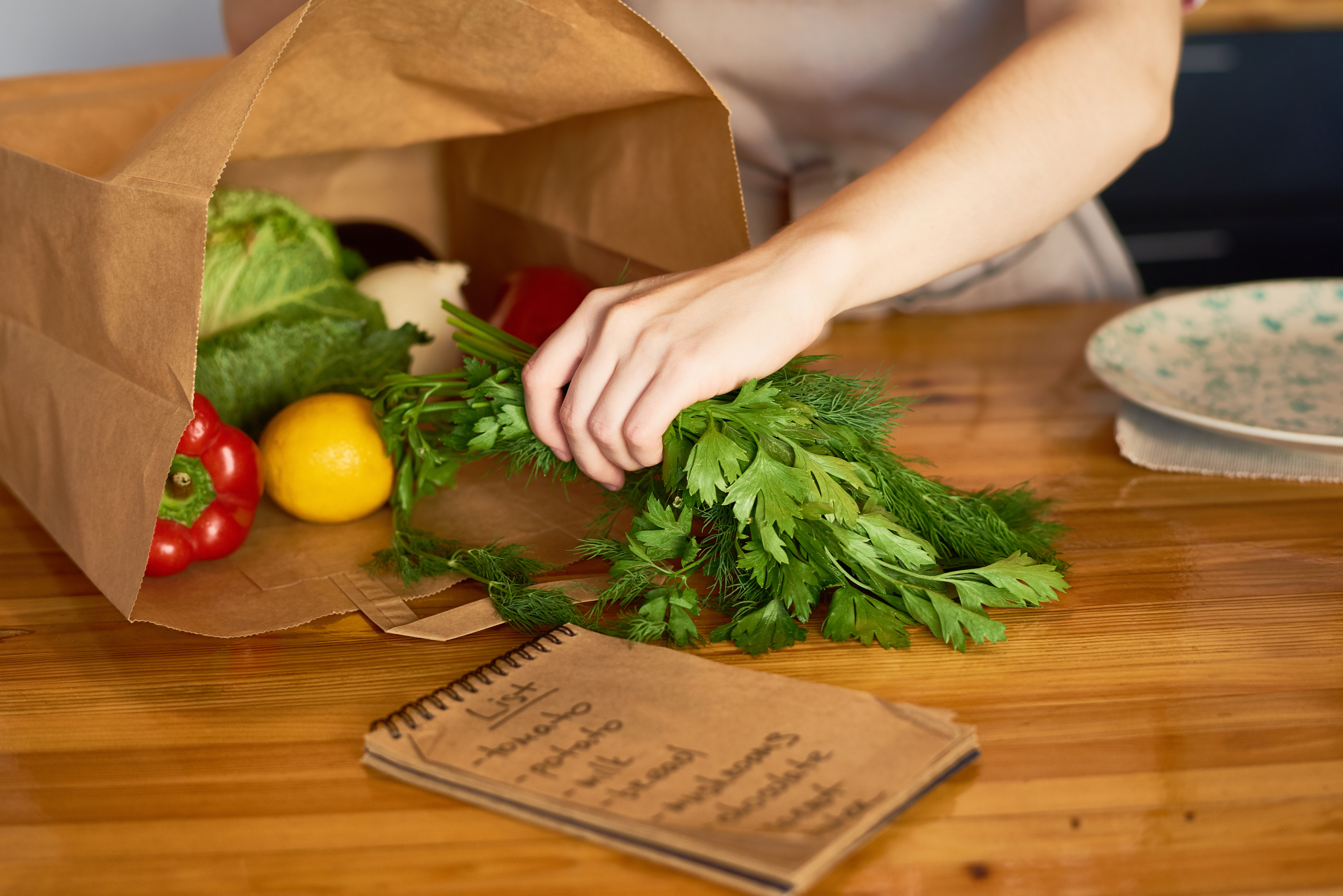 Person unpacking fresh herbs and vegetables with a notebook on a wooden table.