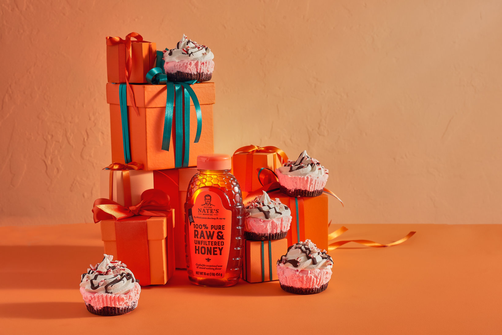 A bottle of Nate's honey, gift boxes and mini peppermint pies on an orange background.