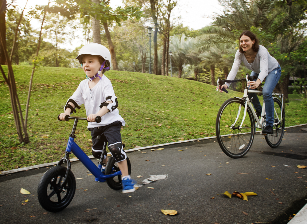 Child on a balance bike and woman on bicycle on a park path.
