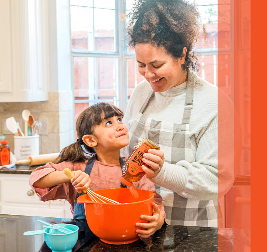 An adult and child smile while cooking together in a kitchen holding a whisk and Nate's honey.