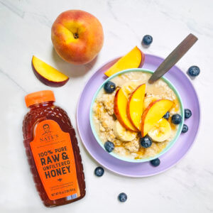 Oatmeal bowl with peach slices and blueberries, beside a bottle of Nate's honey and a whole peach on marble.