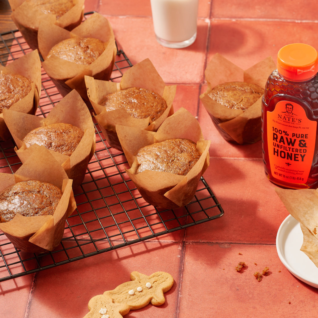 Gingerbread muffins, gingerbread cookies, a bottle of Nate’s honey and glasses of milk on a terracotta surface.