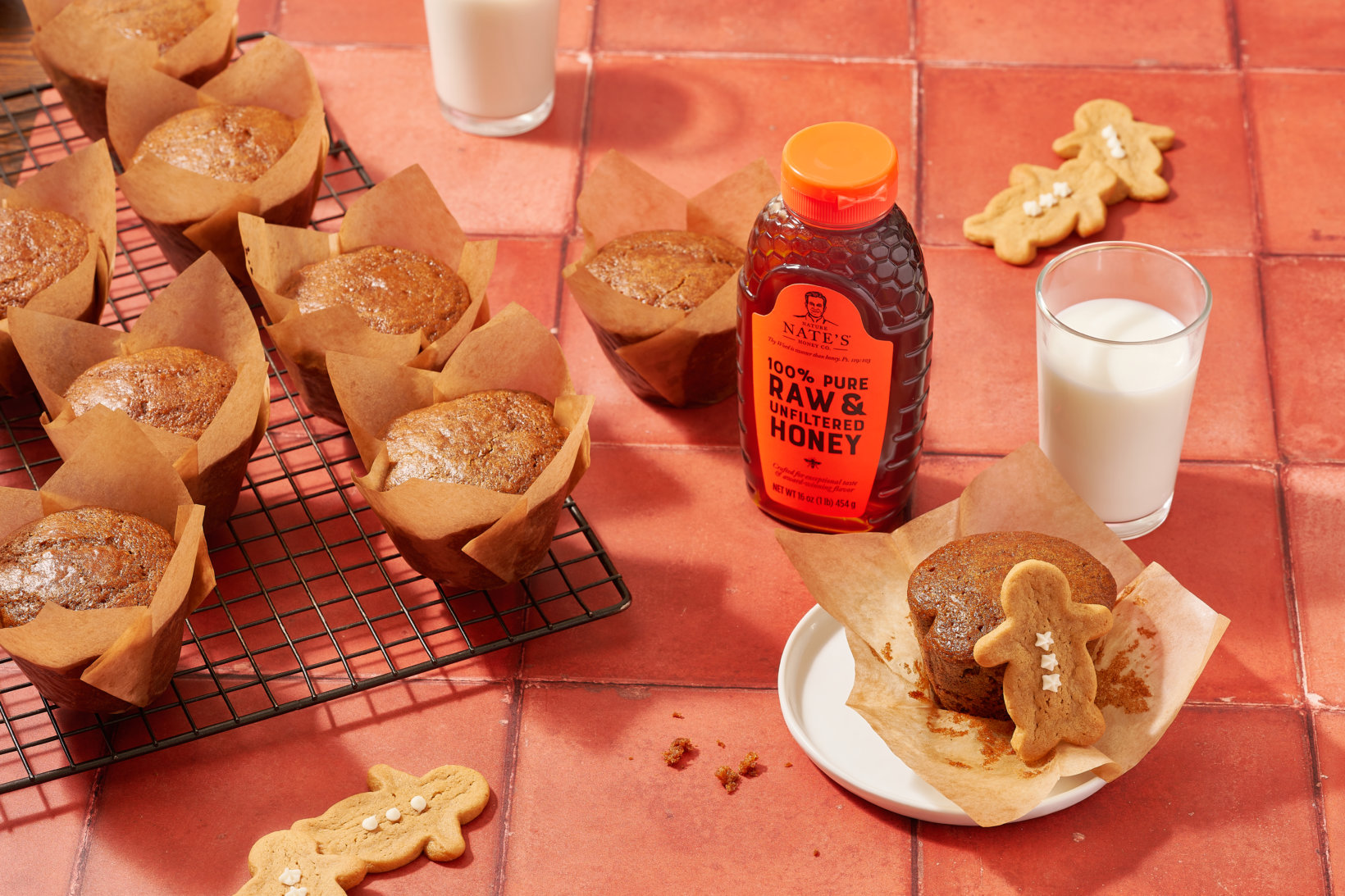 Gingerbread muffins, gingerbread cookies, a bottle of Nate’s honey  and glasses of milk on a terracotta surface.
