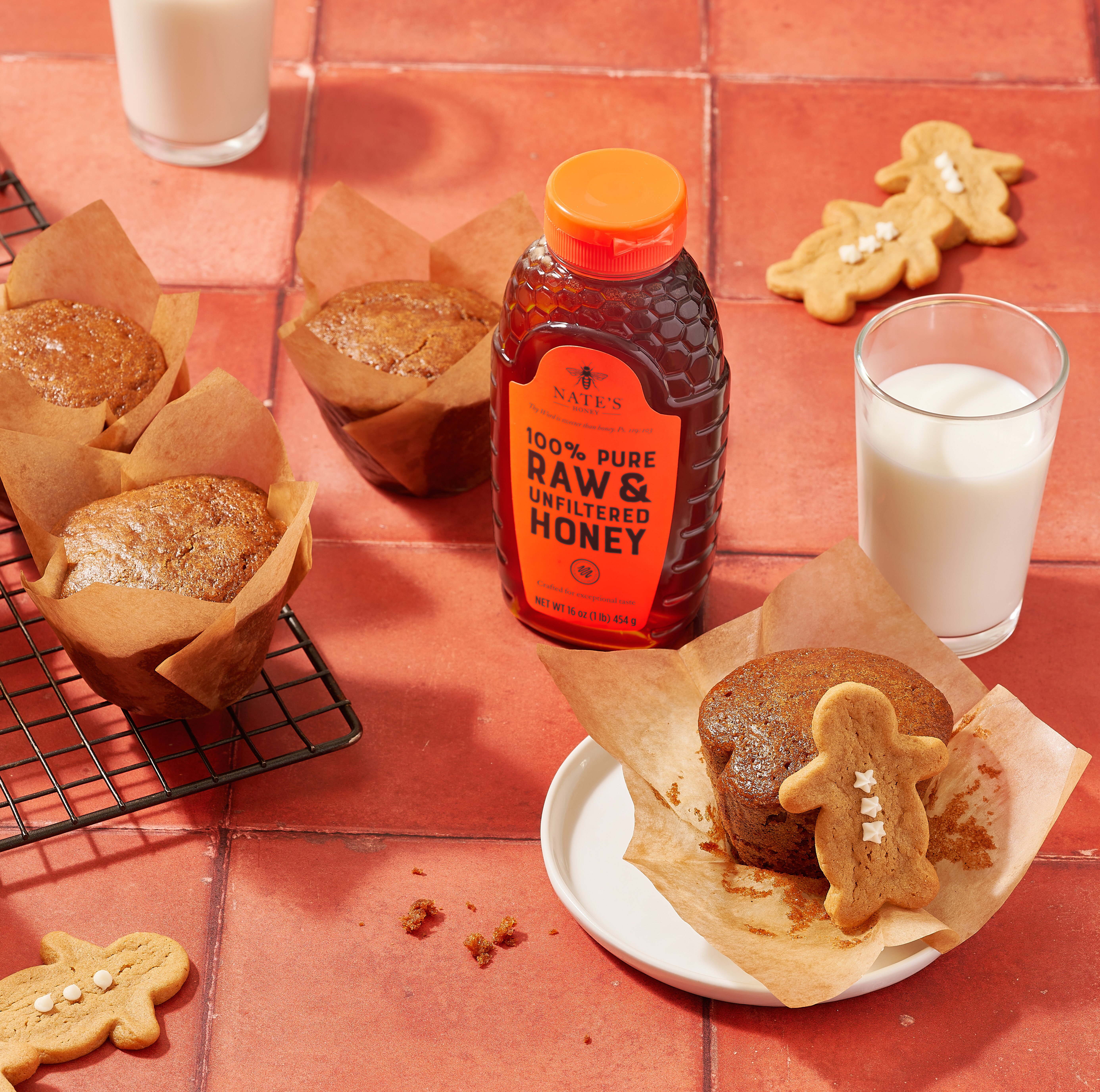 Gingerbread muffins, gingerbread cookies, a bottle of Nate’s honey  and glasses of milk on a terracotta surface.