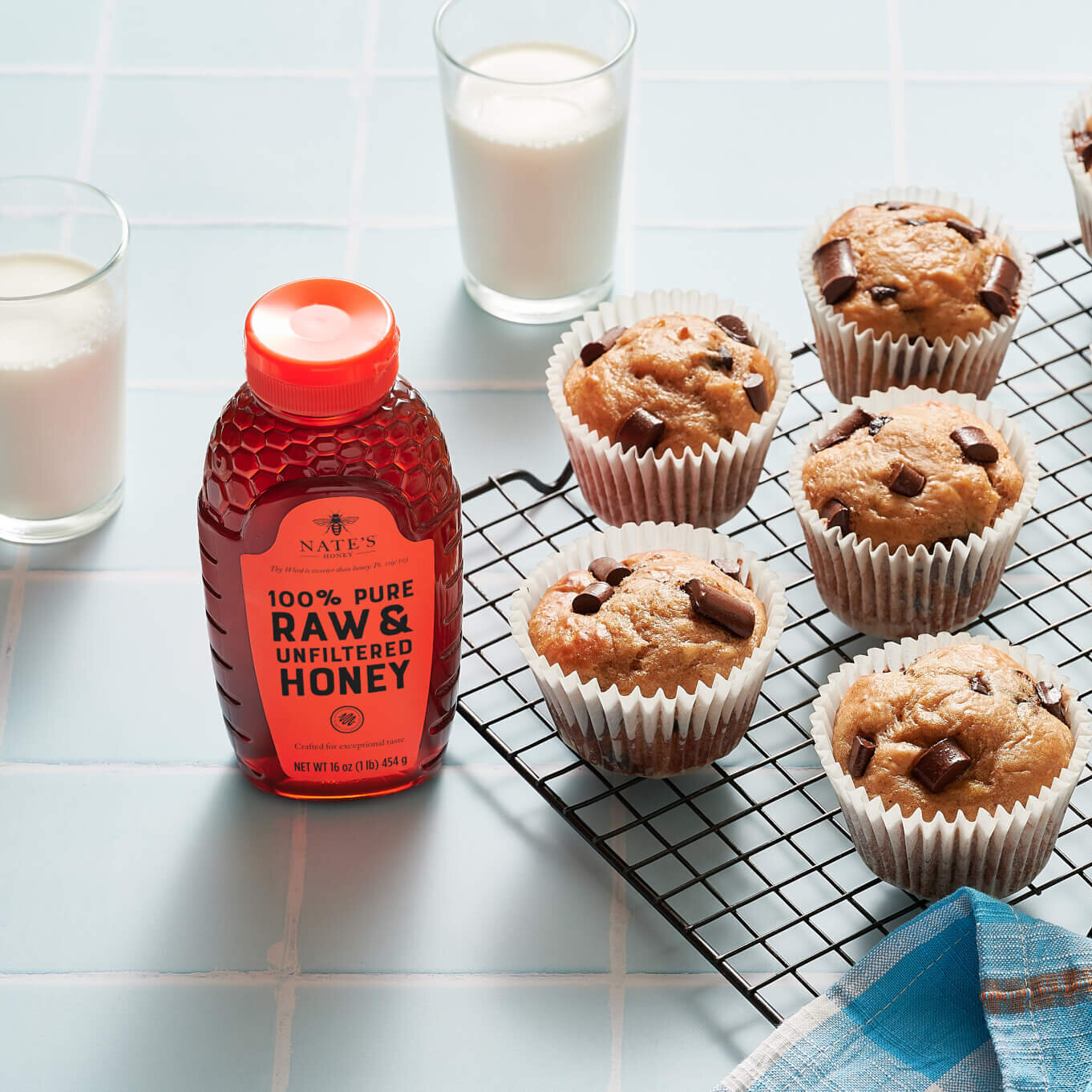 Muffins on a cooling rack, a bottle of Nate's Honey and glasses of milk  on a tiled counter with a blue checkered towel.