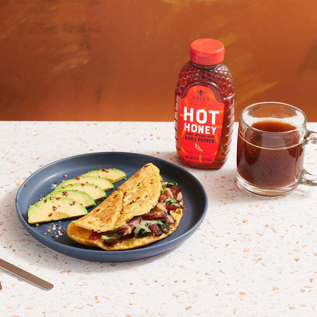 Breakfast set with bacon omelette, avocado, toast, coffee and Nate’s hot honey on an orange surface.