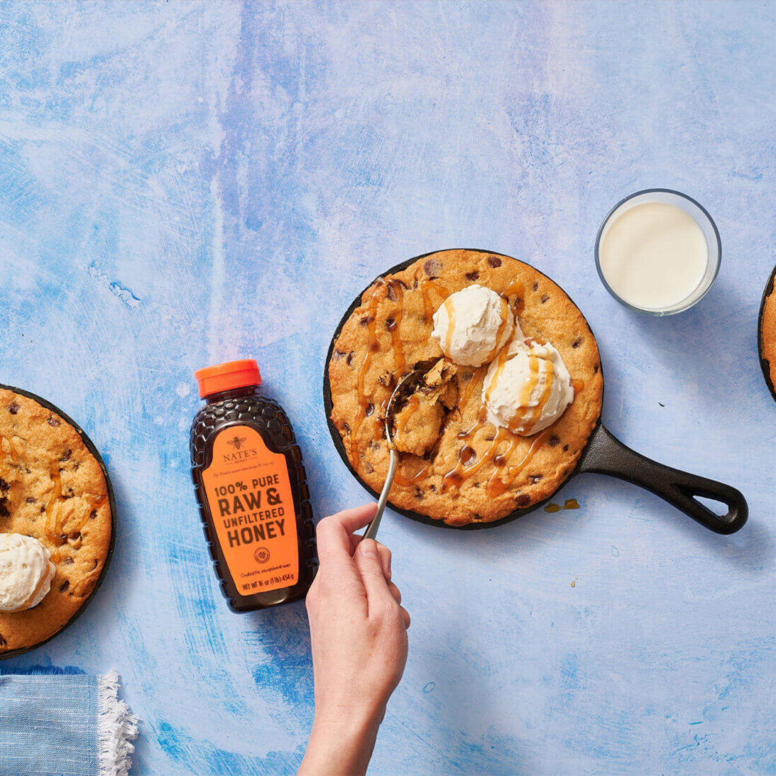 Skillet cookies with ice cream, a bottle of Nate’s honey and glasses of milk with a hand and spoon on a blue background.