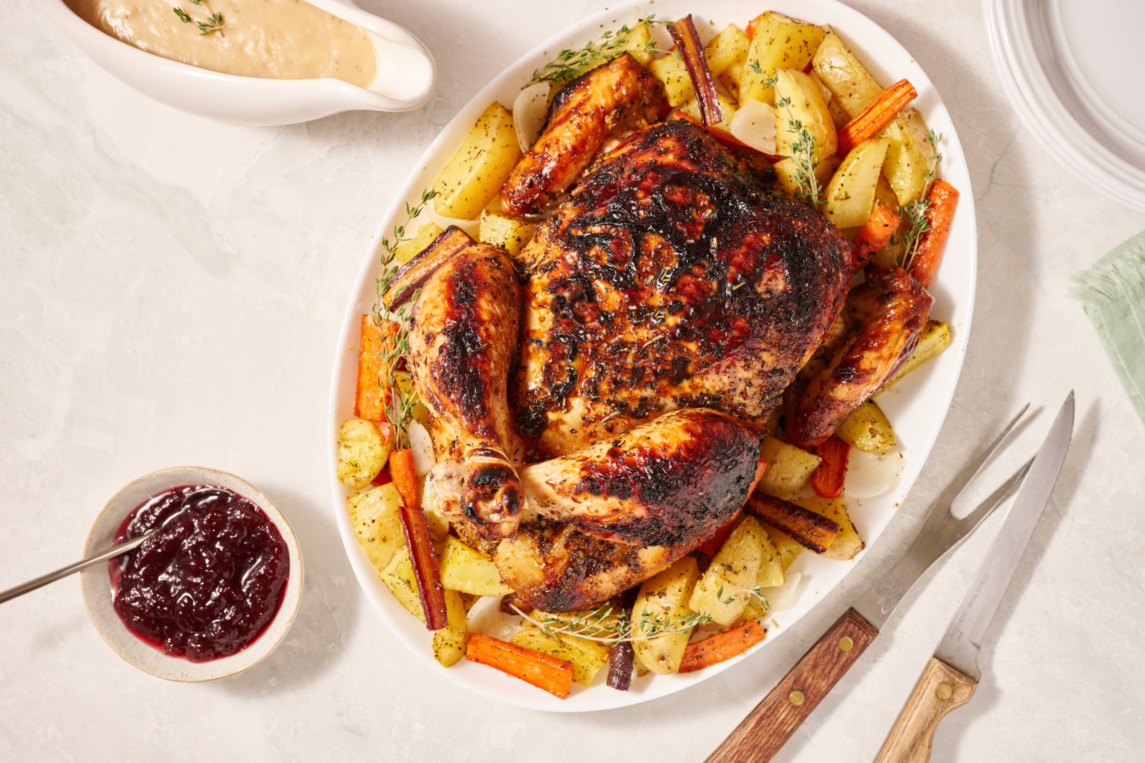 Roasted chicken surrounded by seasoned potatoes and carrots, served with cranberry sauce and gravy, on a marble table.