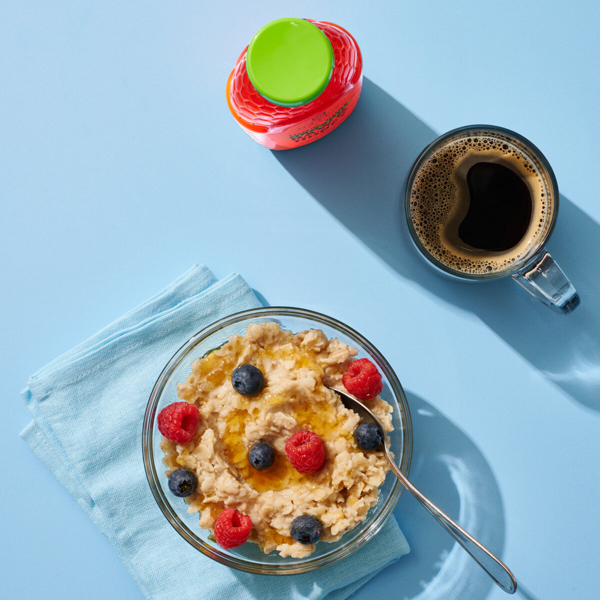 Bowl of oatmeal topped with berries and honey beside a cup of coffee on a light blue surface.