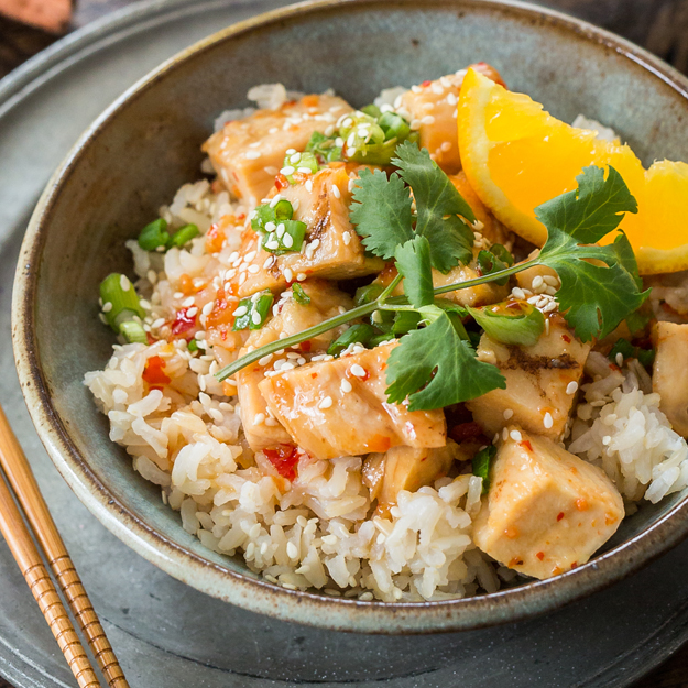 Orange chicken over rice with an orange slice, green onions and cilantro in a ceramic bowl.