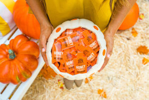 A person in a yellow outfit holds a white pumpkin basket full of Nate’s honey minis, surrounded by large pumpkins.