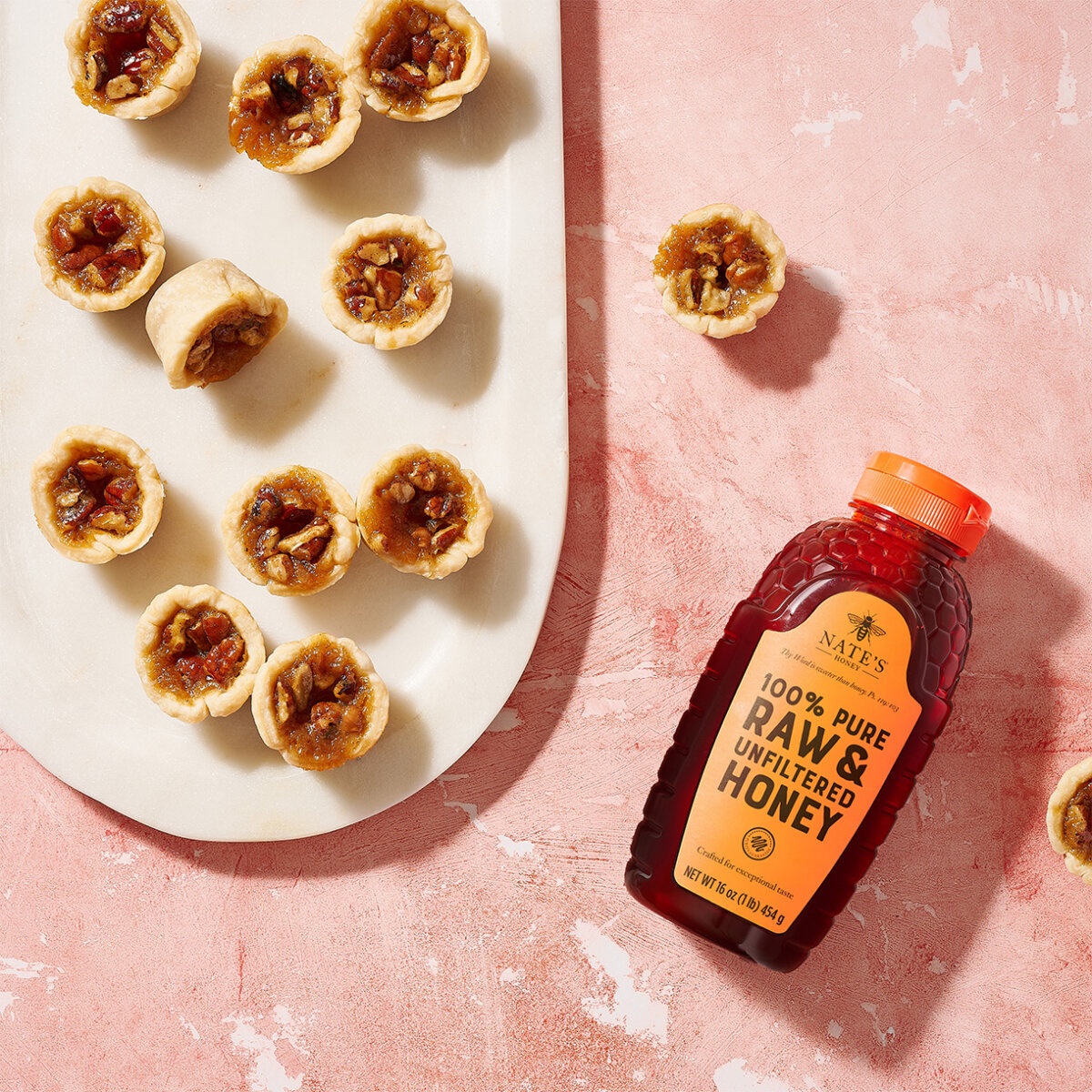 Mini pecan tarts on an oval plate and a bottle of Nate’s Honey on a textured pink surface.