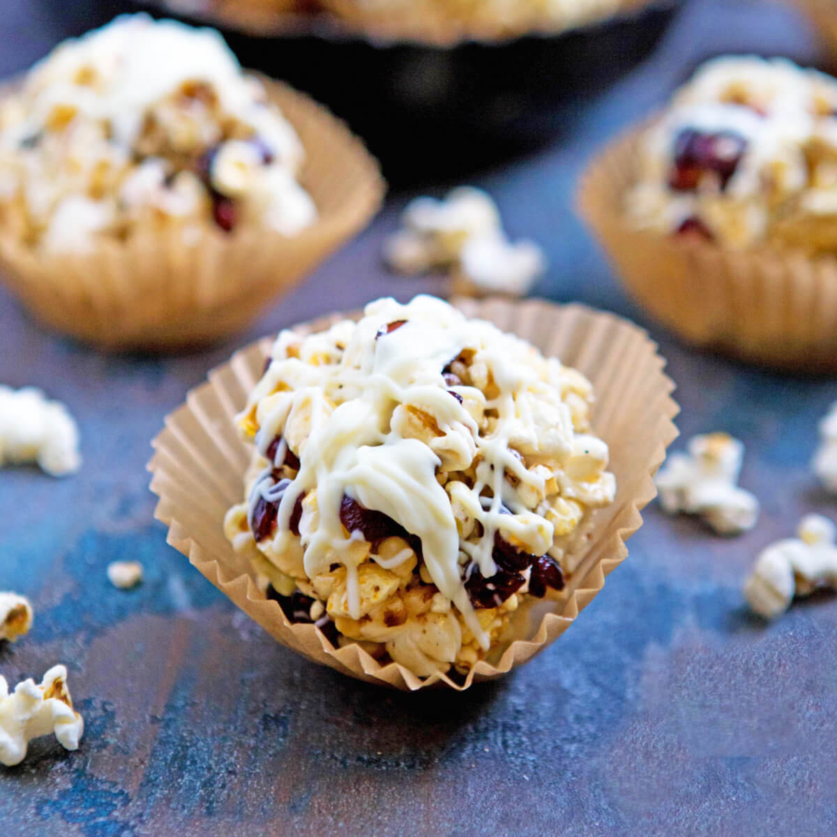 Popcorn balls with cranberries, nuts and white chocolate drizzle in paper cups on a dark surface.