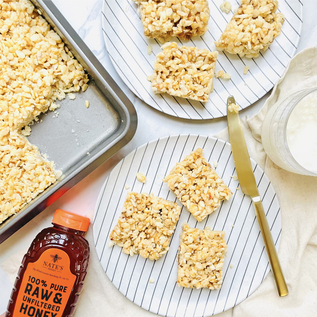 Rice crispy treats made with honey on a striped plate, a bottle of Nate’s Honey, and a glass of milk on marble.