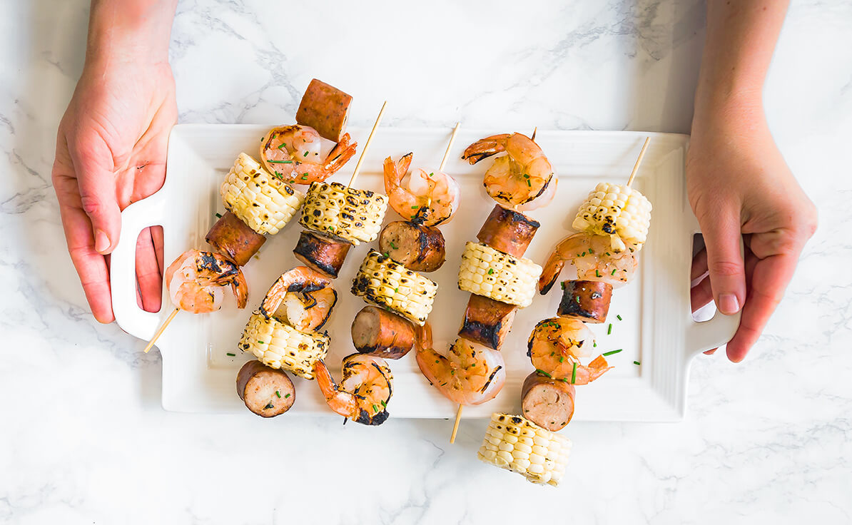 Hands presenting a plate of grilled shrimp and sausage skewers with corn on marble.