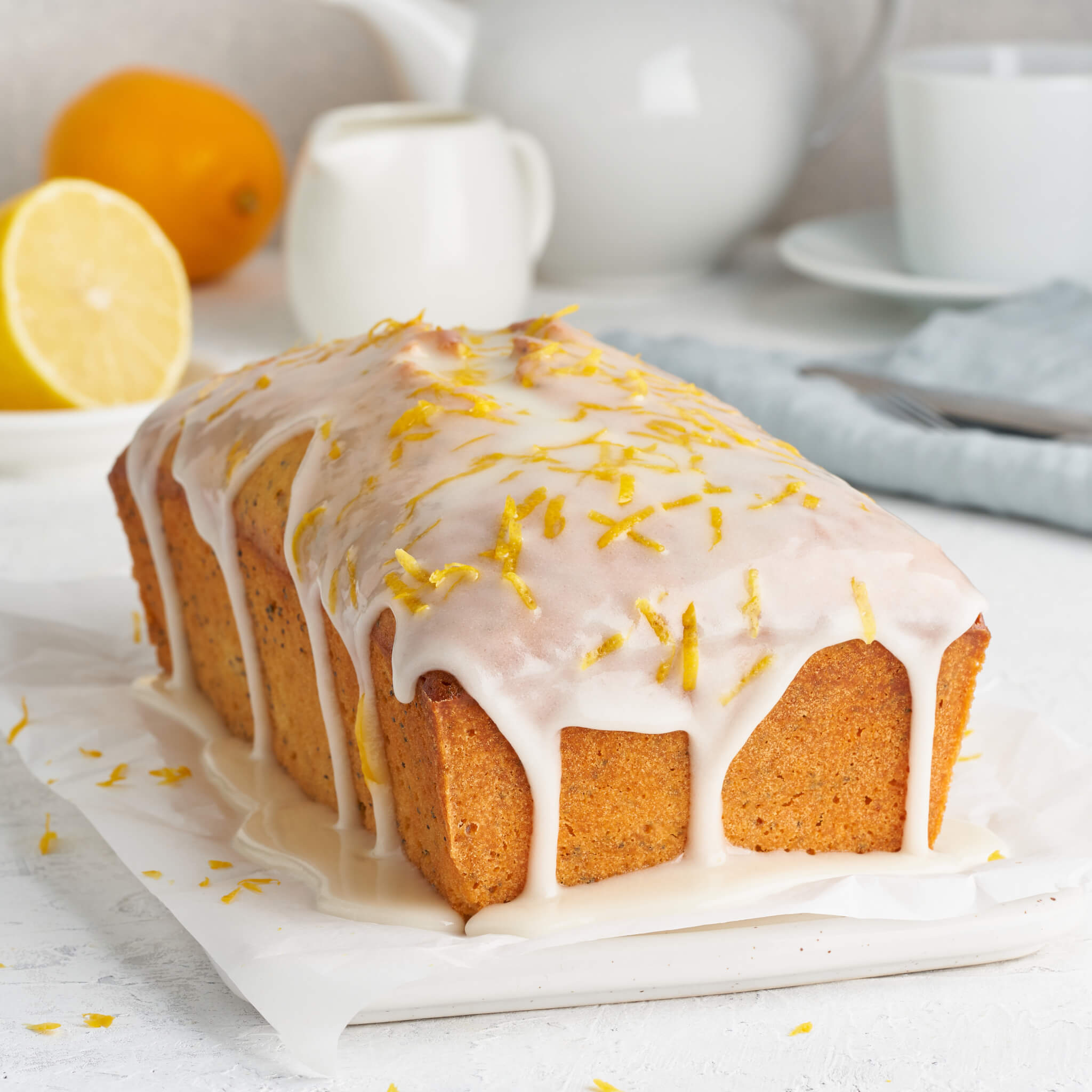 Pound cake made with honey and topped with white icing and lemon zest on a white table, kitchen items in the backdrop. 