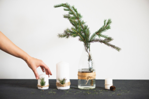 Hand reaching for a candle beside a vase with a pine branch on a black table, white background.