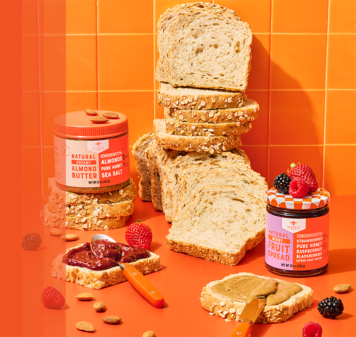 Nate’s Honey almond butter and berry fruit spread displayed with sliced bread, toast topped with fruit spread and almond butter, fresh berries and almonds on a bright orange background.