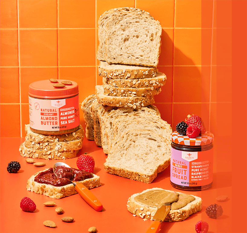 Nate’s Honey almond butter and berry fruit spread displayed with sliced bread, toast topped with fruit spread and almond butter, fresh berries, and almonds on a bright orange background.