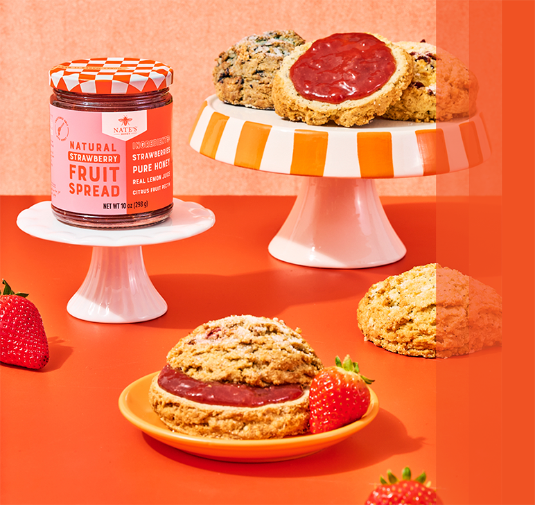 Jar of Nate’s Natural Strawberry Fruit Spread displayed with scones topped with strawberry spread and fresh strawberries on an orange background.