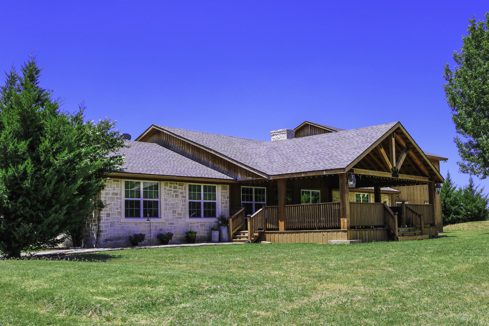 A single-story stone house with a prominent wooden gable, front deck, and green lawn, under a clear blue sky. A single-story stone house with a prominent wooden gable, front deck, and green lawn, under a clear blue sky.