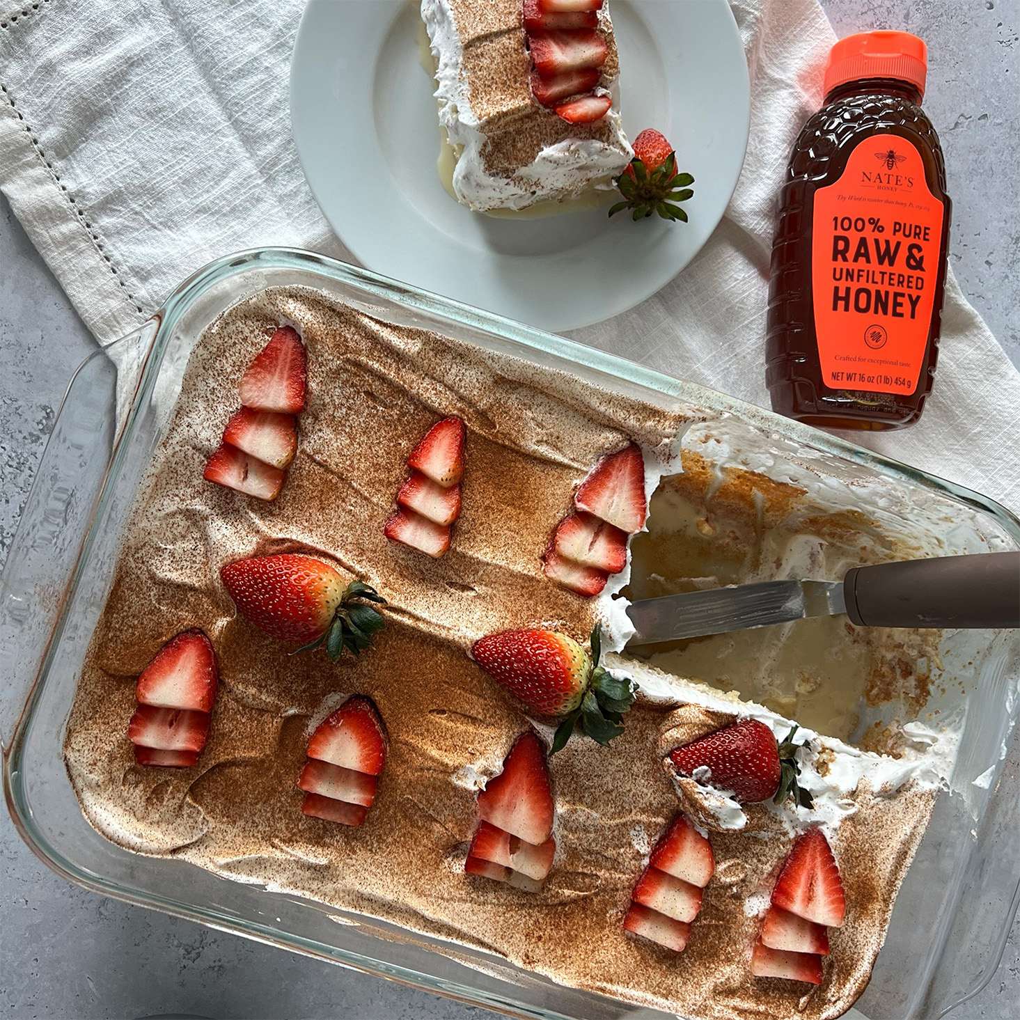 Tres leches cake with white frosting and garnished with strawberries and cocoa powder, next to a bottle of Nate's Honey.