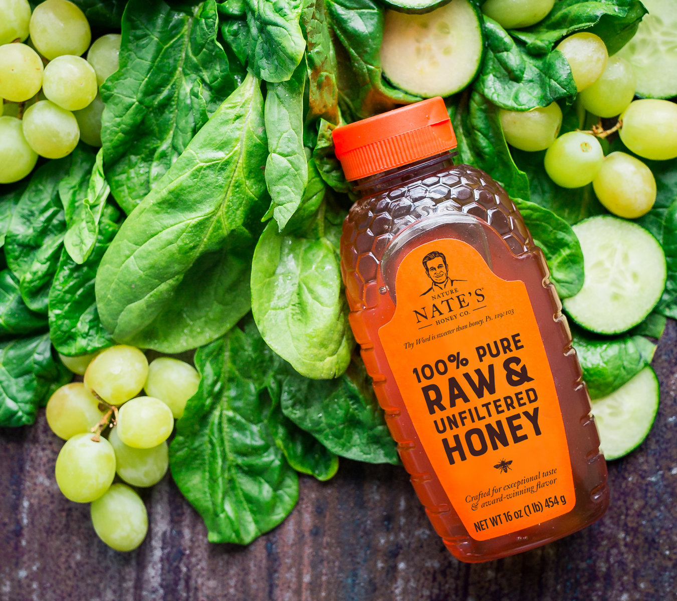 A bottle of Nate's honey on spinach, with grapes and cucumbers around.